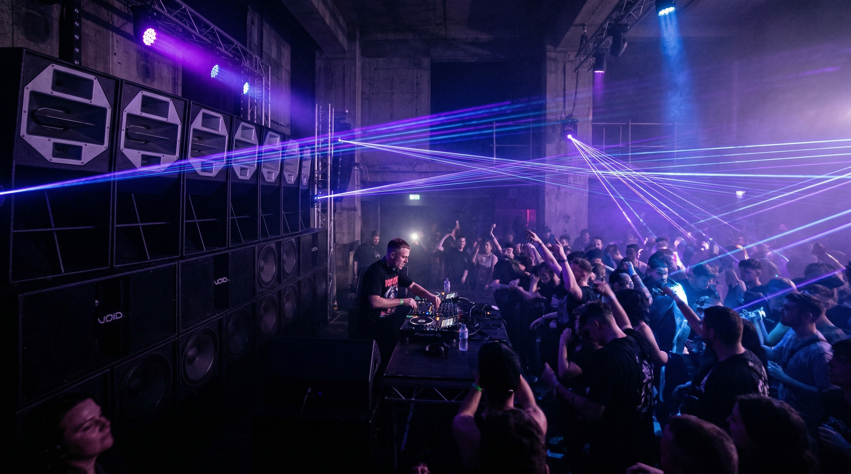 Intense bass music event with purple and blue lasers cutting through fog and speakers stacked wall-to-wall